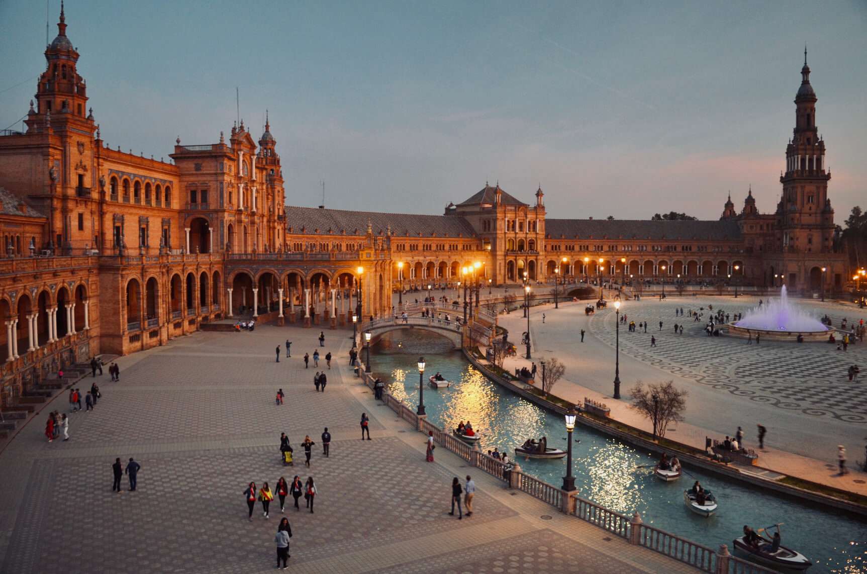 Der Platz zeigt beeindruckende Architektur, Wasserspiele und ein reges Treiben von Menschen bei Dämmerung.