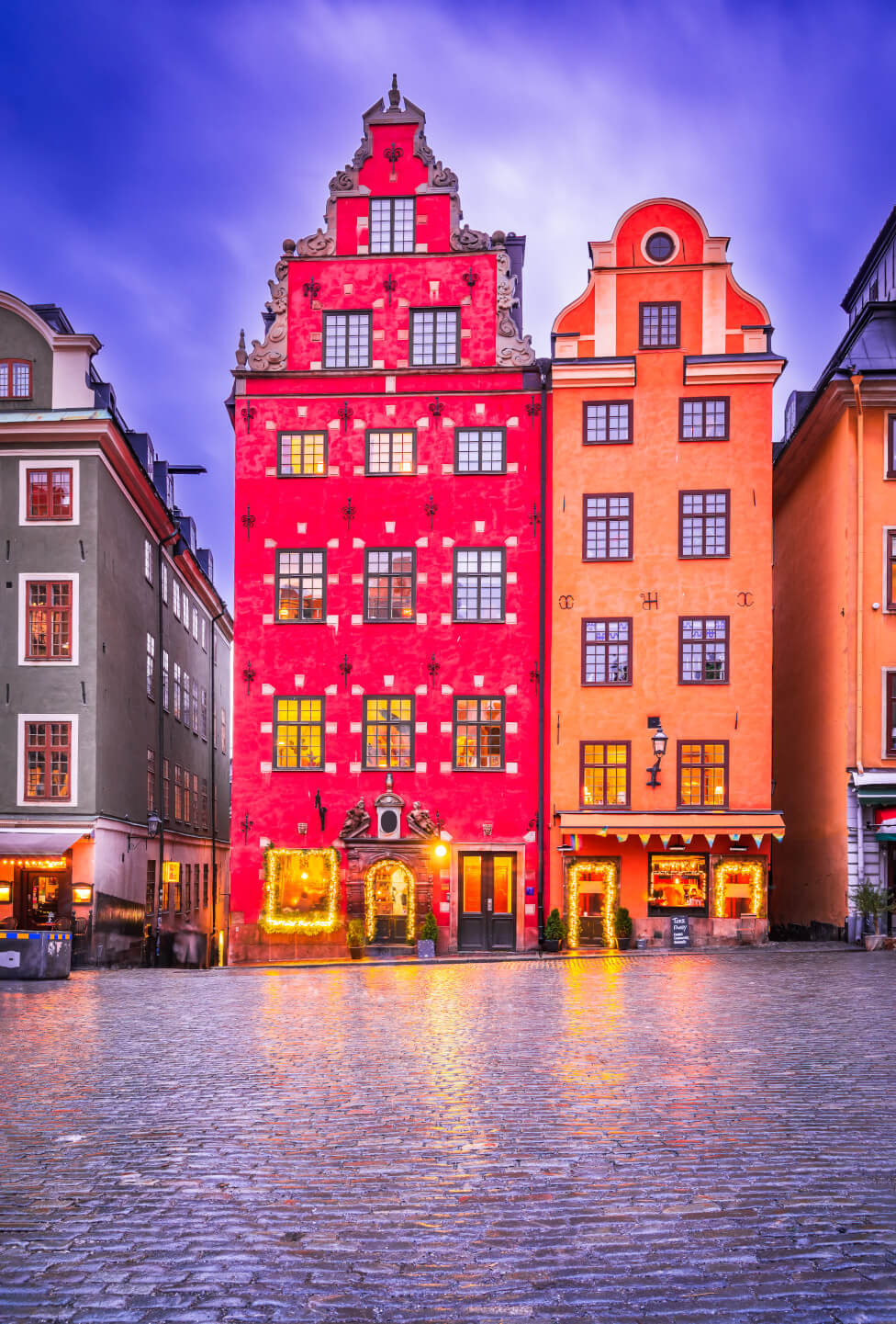 Bunte historische Gebäude stehen an einem Platz, mit festlicher Beleuchtung und reflektierendem Pflaster.