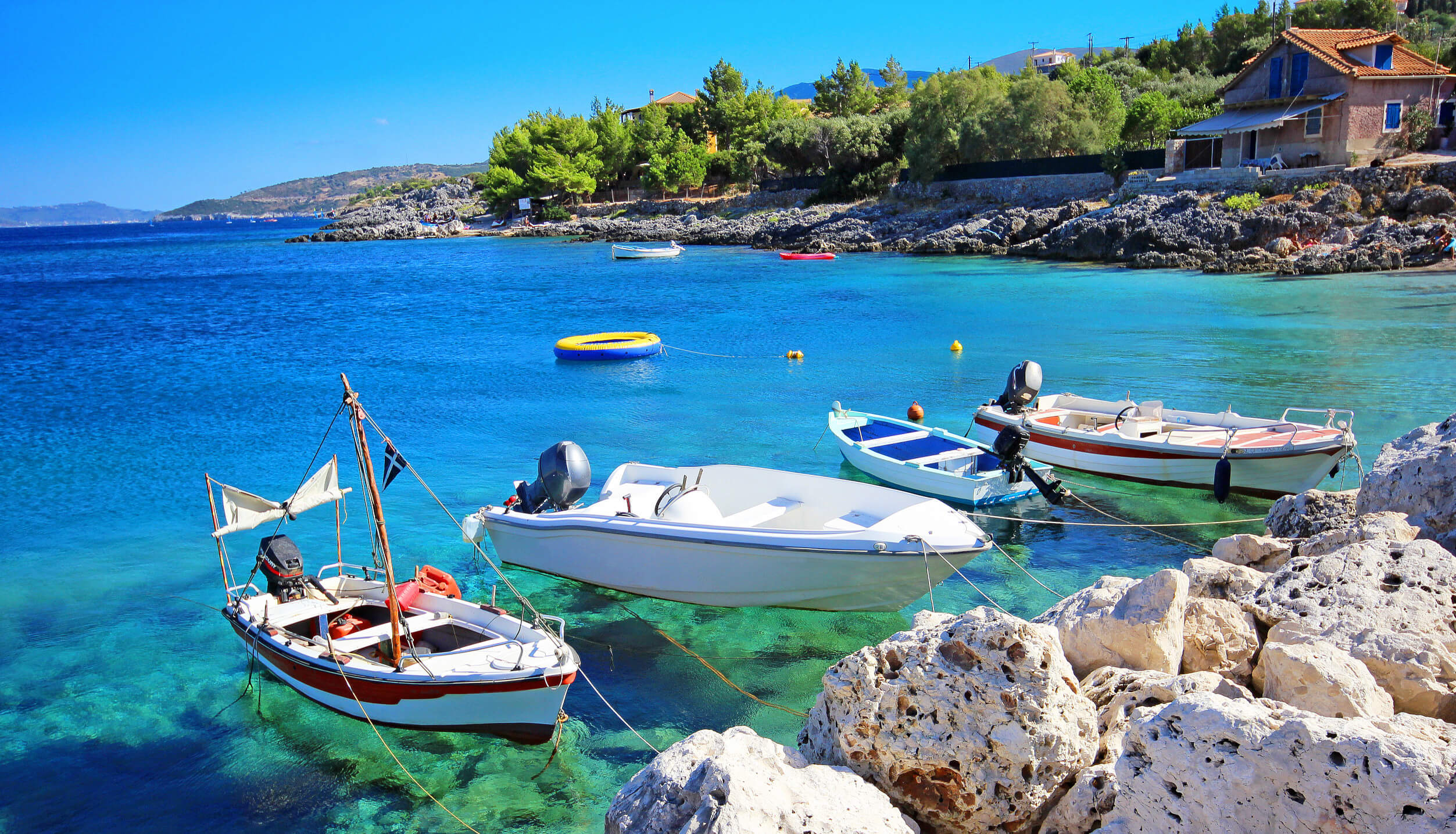 Schöne Küstenlandschaft mit Booten im klaren Wasser, umgeben von Felsen und grünen Bäumen unter blauem Himmel.