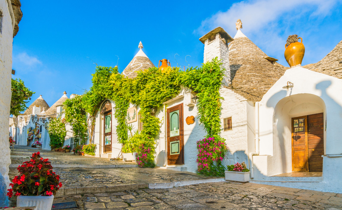 Bunte Blumen und grüne Pflanzen umgeben charmante weiße Häuser mit charakteristischen Dächern im Sonnenlicht.