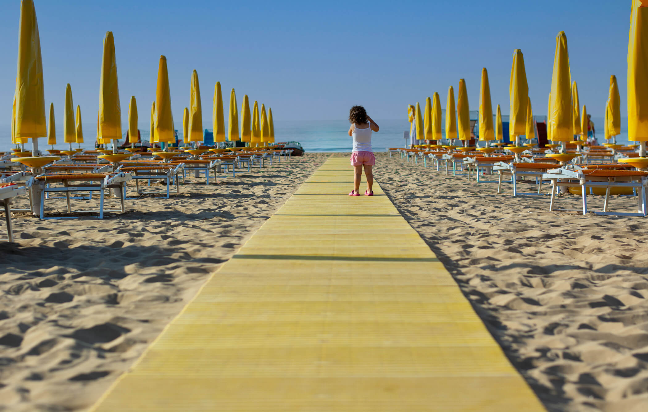 Ein Mädchen läuft auf einem gelben Weg am Strand, umgeben von aufgestellten Sonnenschirmen und Liegen.