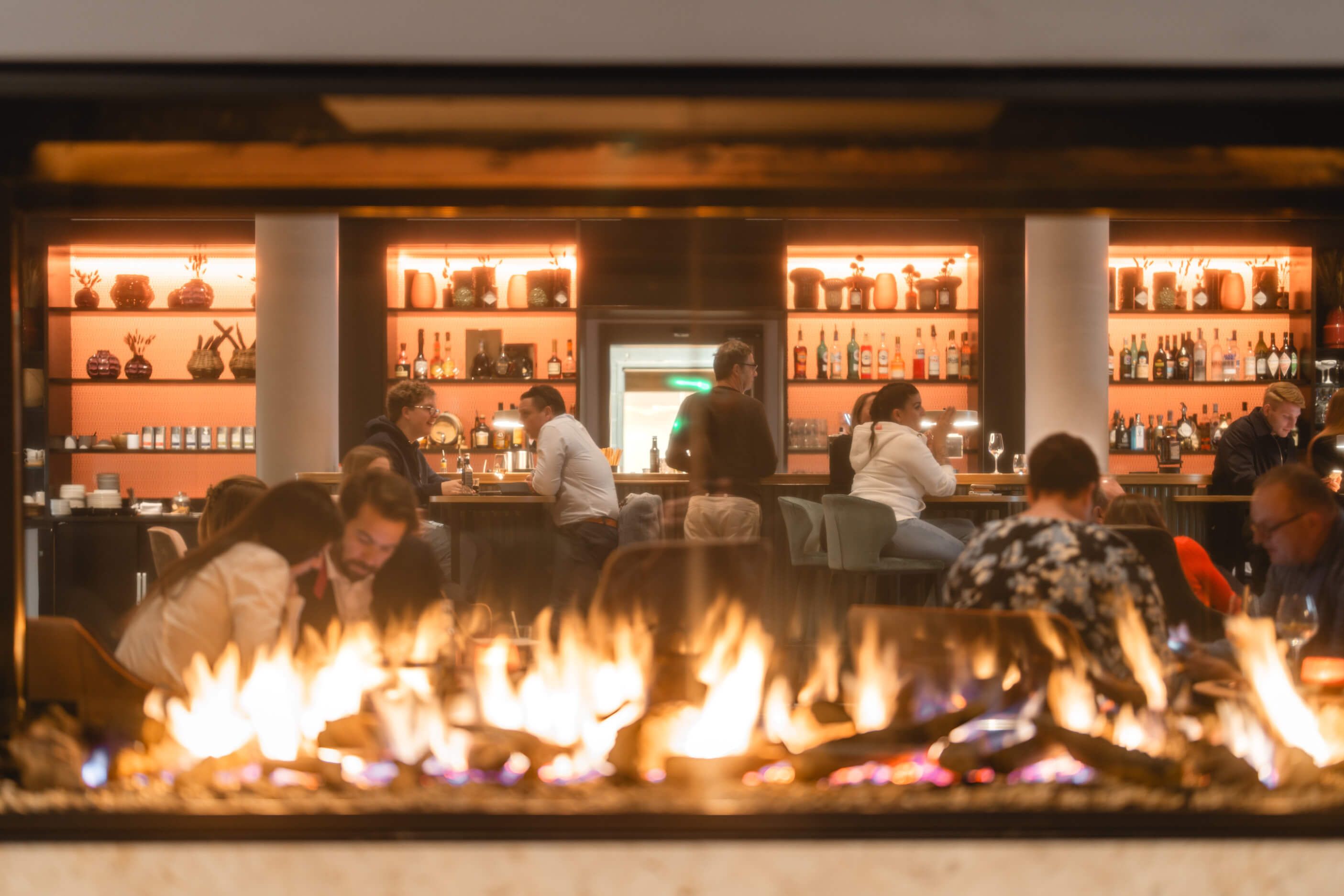Ein modernes Restaurant mit einem Kamin im Vordergrund und Gästen, die an der Bar und Tischen sitzen.