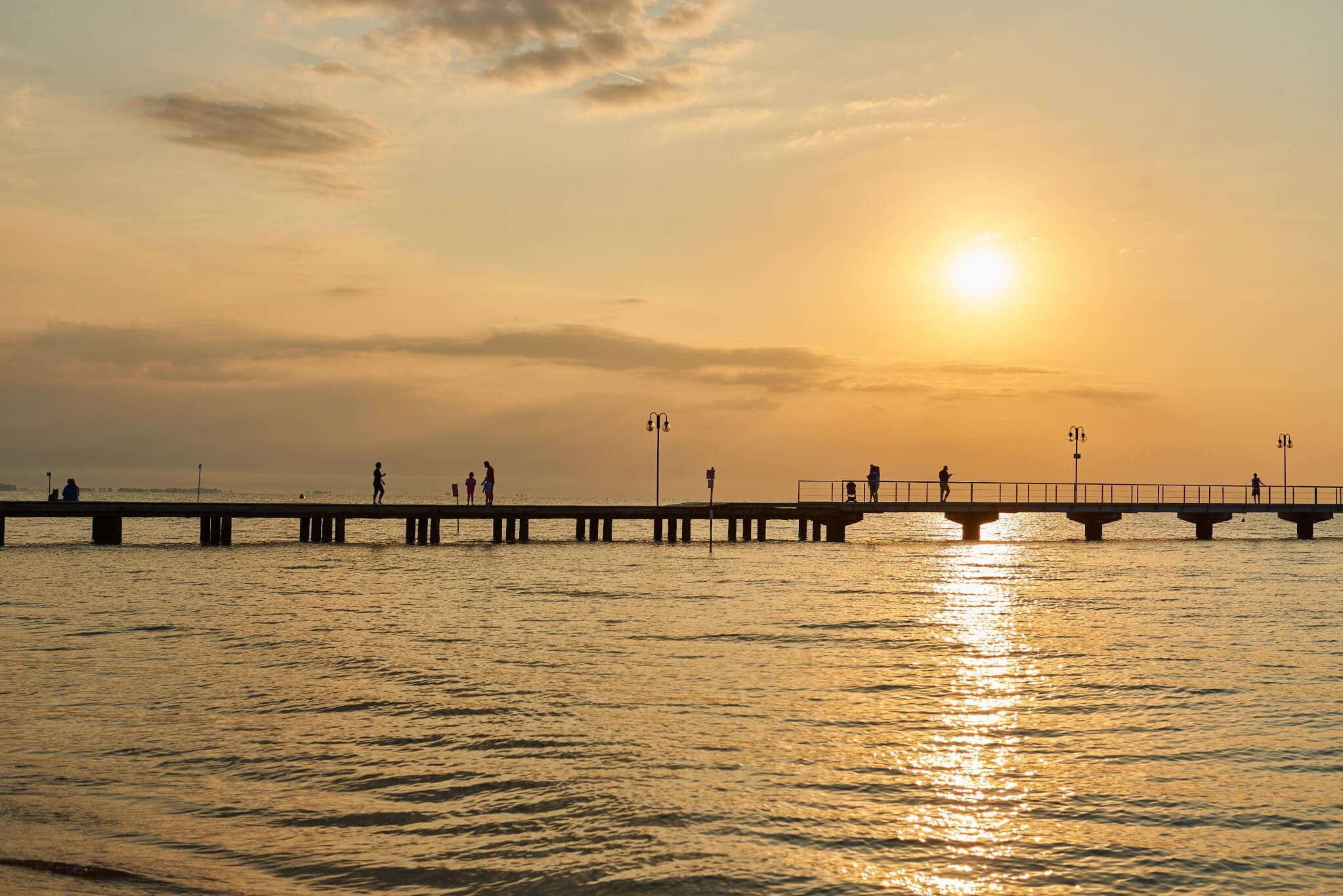 Ein Pier bei Sonnenuntergang, Menschen spazieren entlang des Wassers, drapierte Wolken am Himmel.