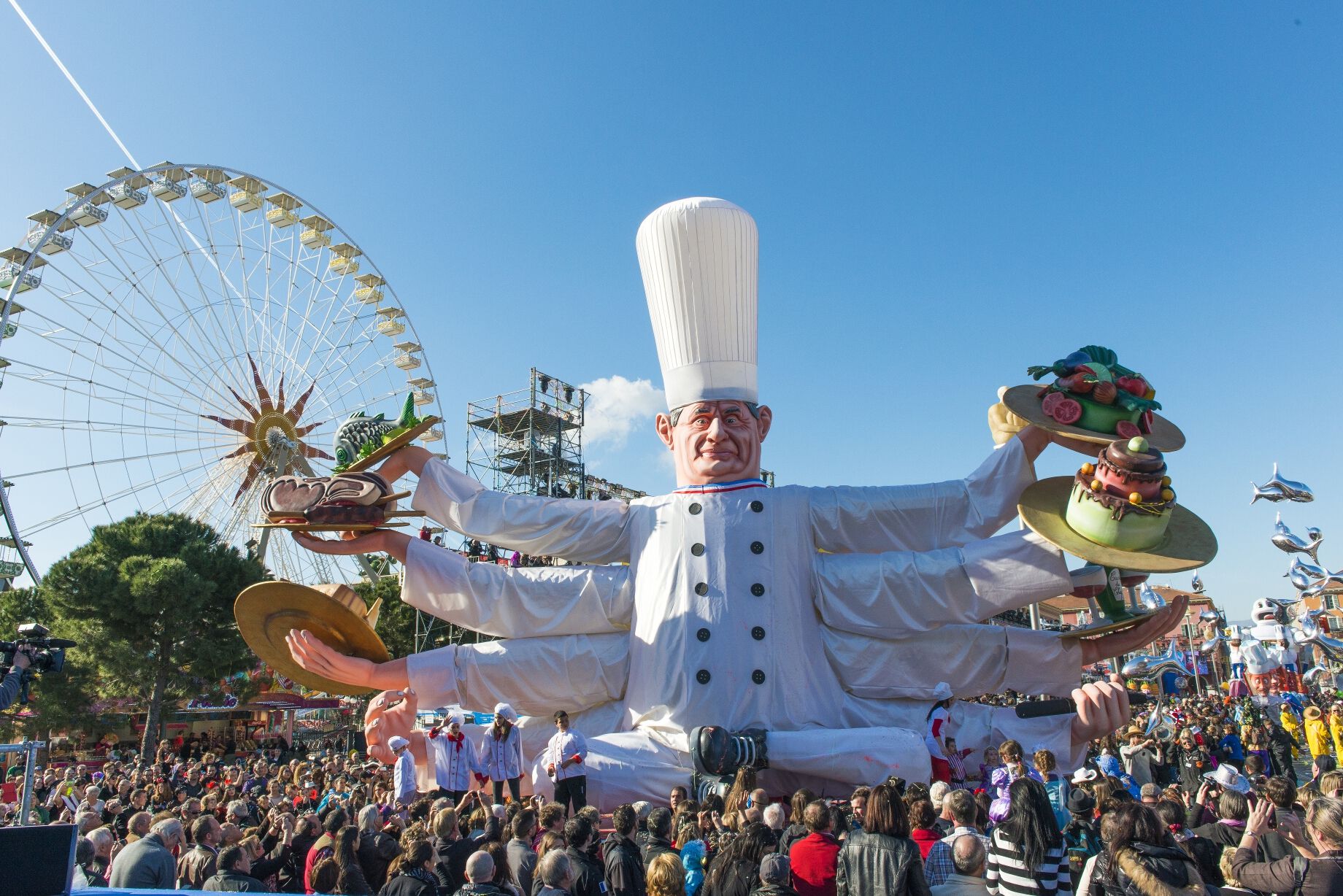 Ein großer Koch mit vielen Armen präsentiert bunte Essen, umgeben von Zuschauern und einem Riesenrad im Hintergrund.