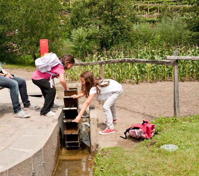 Zwei Mädchen spielen an einem Wasserspiel, während ein Erwachsener in der Nähe sitzt. Grüne Landschaft im Hintergrund.