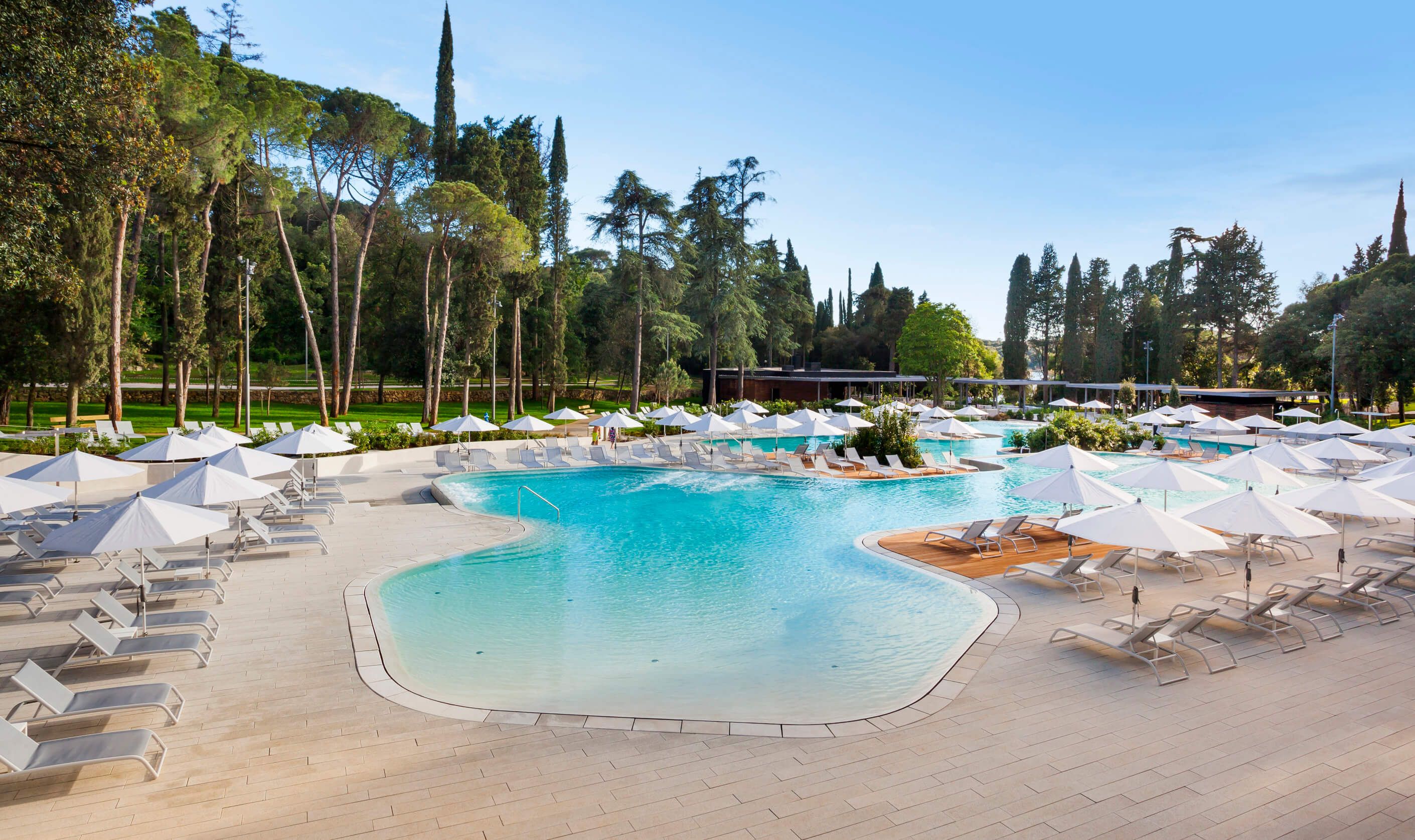 Ein eleganter Poolbereich mit Liegen und Sonnenschirmen, umgeben von grünen Bäumen und blauem Himmel.