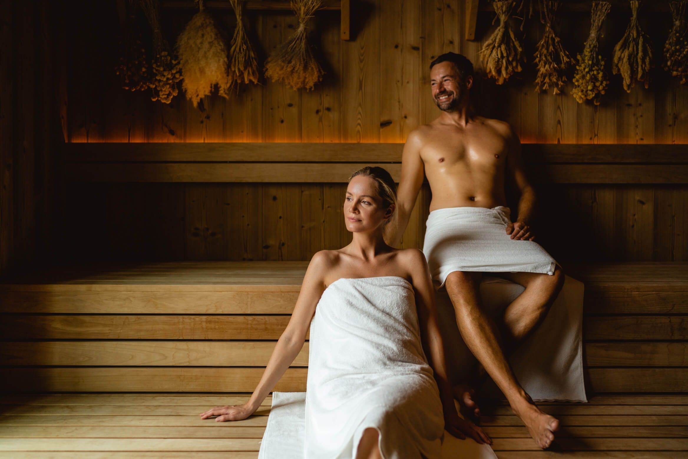 Ein Mann und eine Frau entspannen in einer Sauna, beide mit Handtüchern bekleidet, in einer gemütlichen Holzumgebung.