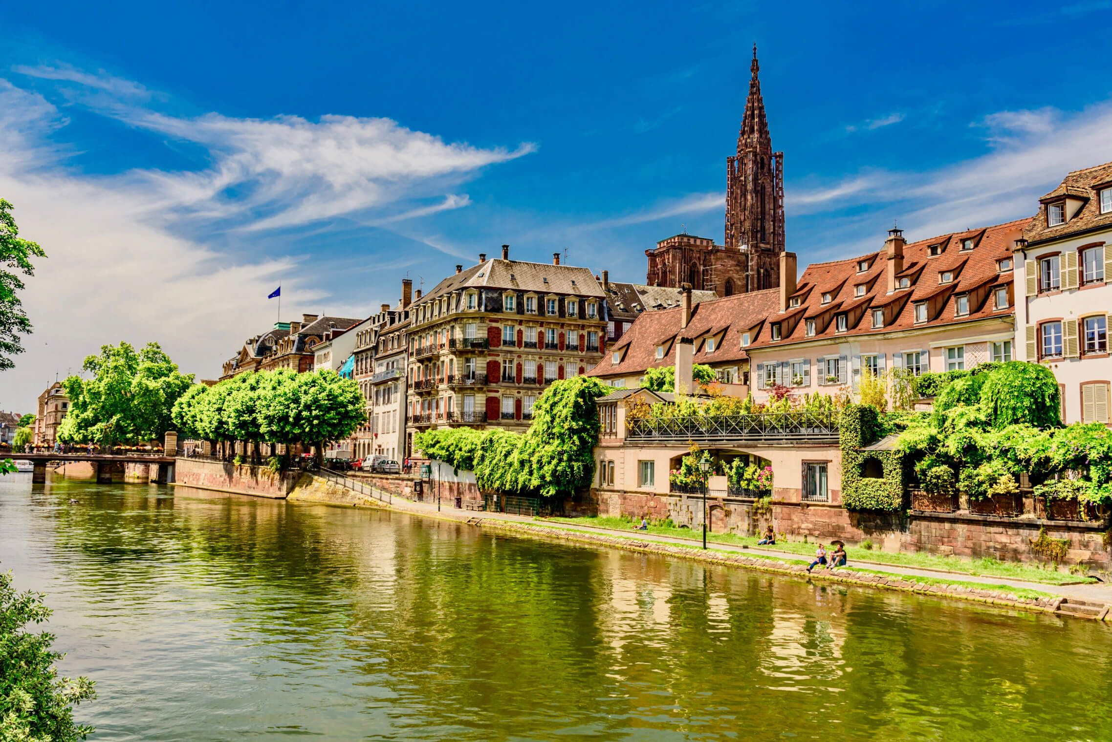 Idyllische alte Gebäude am Flussufer, grünes Laub, strahlend blauer Himmel und der beeindruckende Münster in Straßburg.
