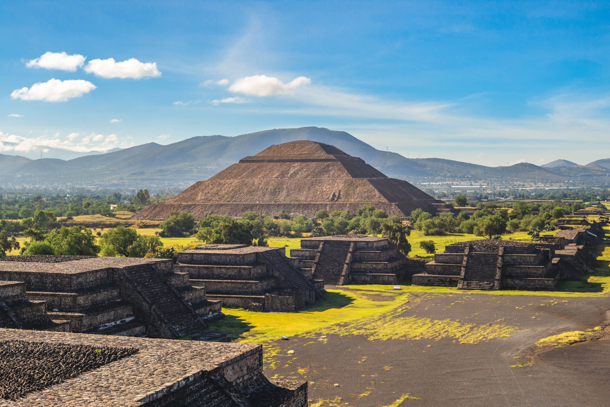 Die Pyramide von Teotihuacan erhebt sich majestätisch über eine grüne Landschaft mit Bergen im Hintergrund.