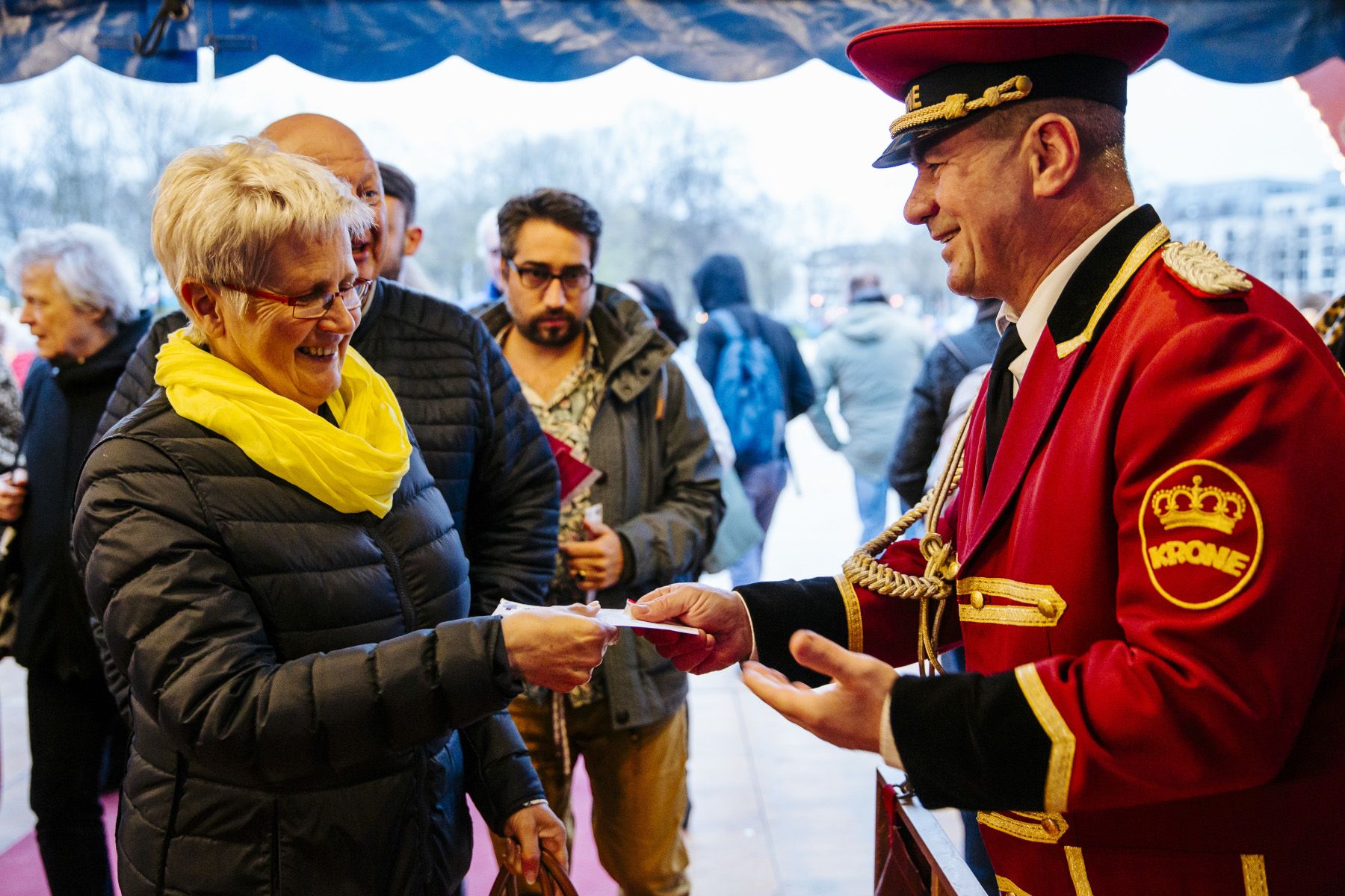 Eine Frau mit gelbem Schal erhält ein Ticket von einem uniformierten Mann in rot. Im Hintergrund warten weitere Gäste.