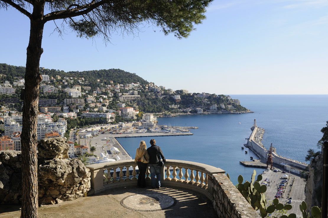 Ein Paar genießt die Aussicht auf die Küste von Nizza mit Hafen und Bergen im Hintergrund.