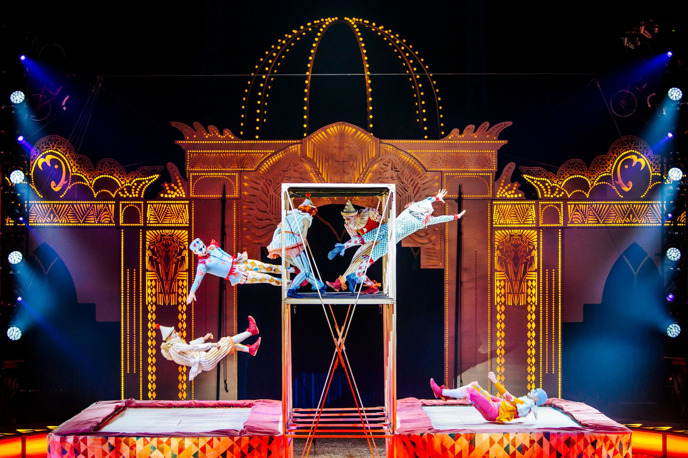 Zwei Clowns springen von einem Trampolin auf eine Bühne mit buntem, leuchtendem Hintergrund. Aufregende Zirkusatmosphäre!