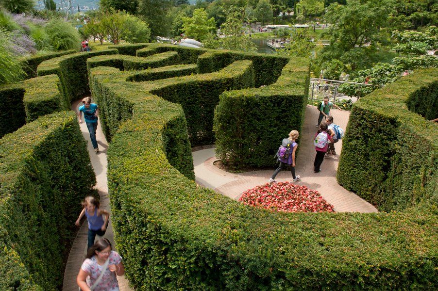 Ein labyrinthartiger Garten mit hohen Hecken, in dem Menschen umherlaufen und Blumen in der Mitte stehen.