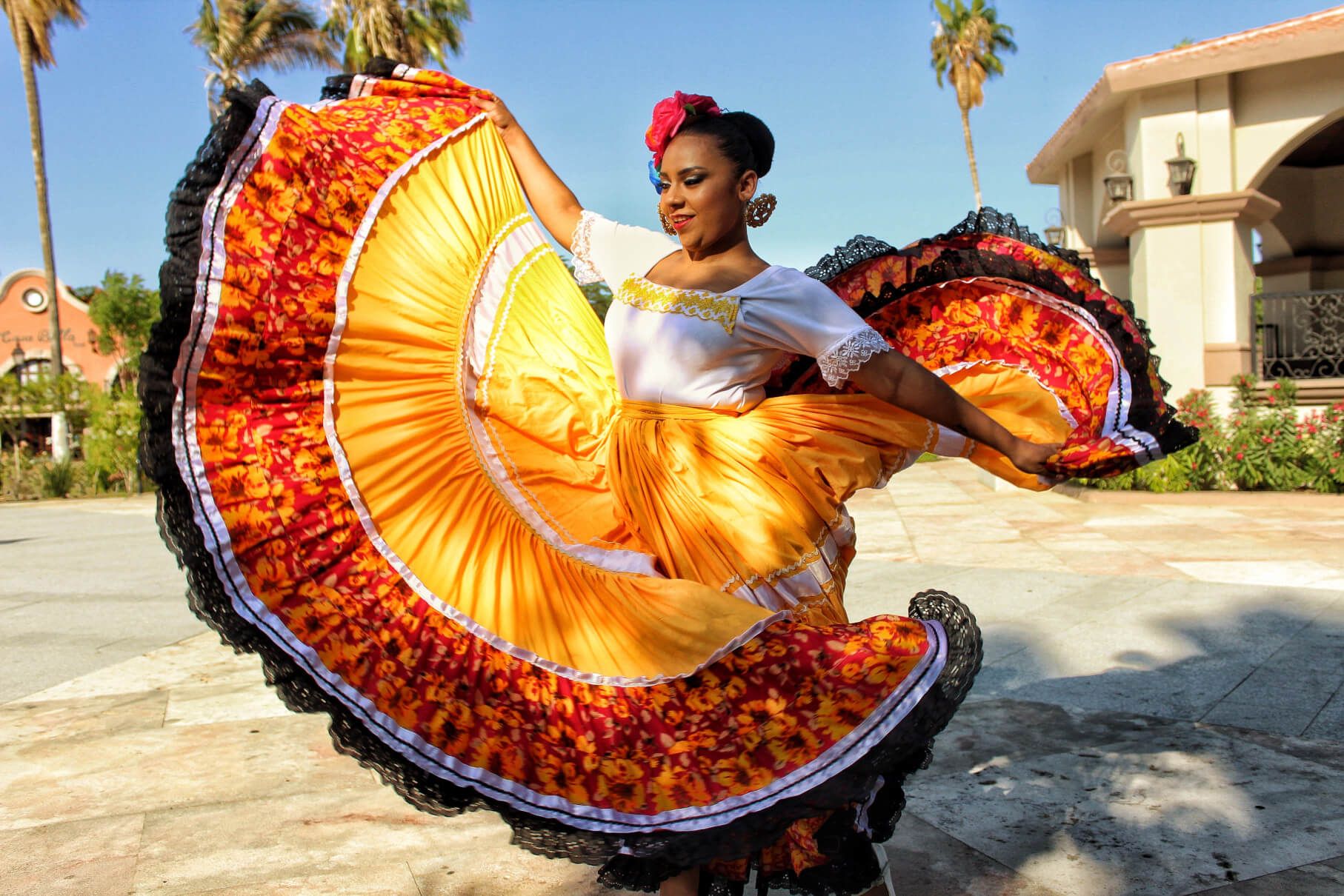 Eine Tänzerin in farbenfrohem Kleid tanzt vor einer Kulisse mit Palmen und sonnigem Himmel.