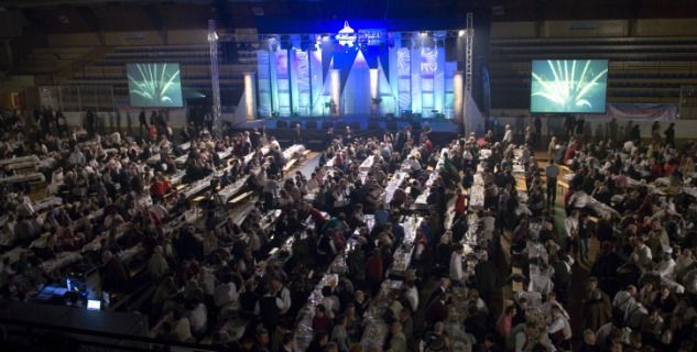 Ein großer Saal voller Menschen bei einer Veranstaltung, mit Tischen und einer Bühne im Hintergrund.