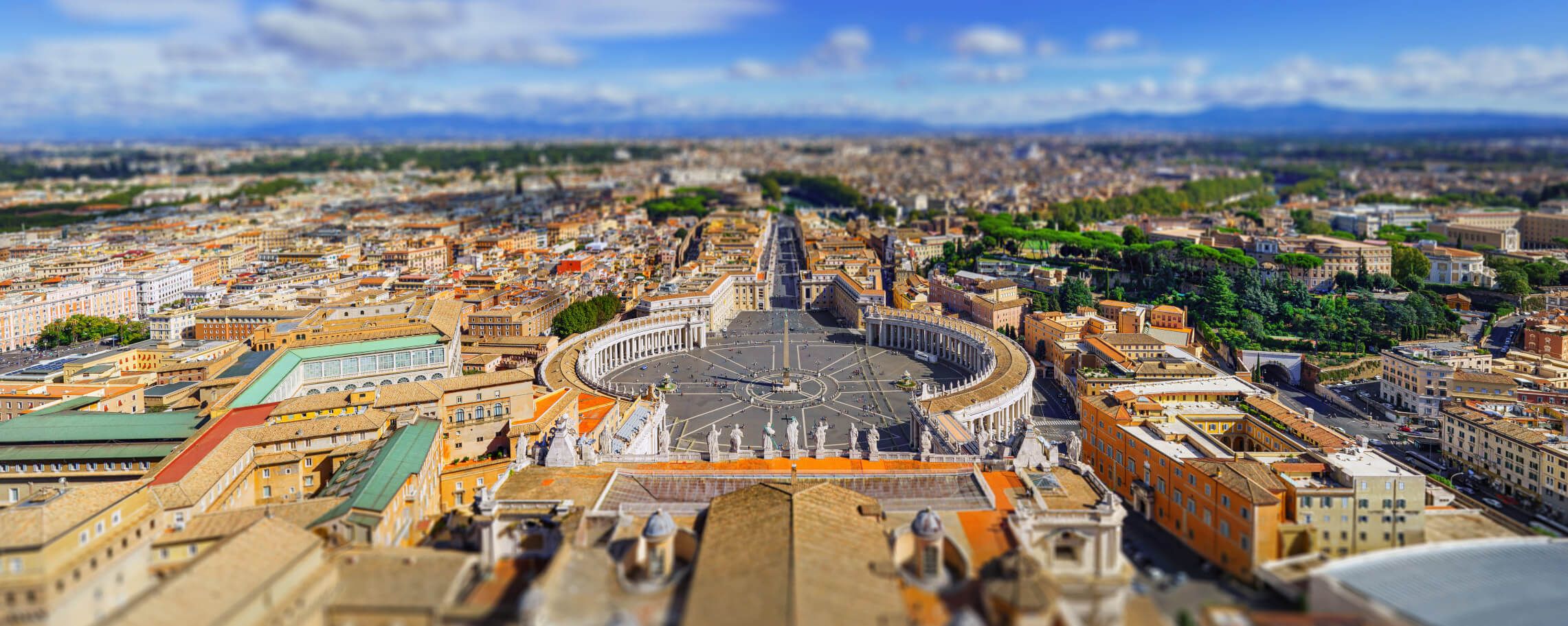 Eine Panoramaaufnahme von Rom, mit dem Petersplatz und historischen Gebäuden im Hintergrund. Schönes Wetter und grüne Bäume.
