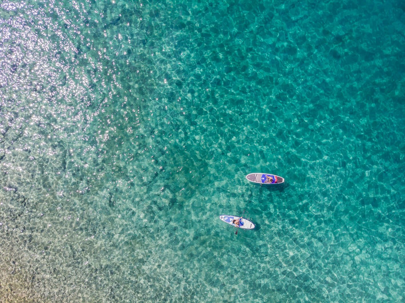 Zwei Stand-up-Paddler gleiten über kristallklares, aquablaues Wasser in der Sonne. Schöne Reflexionen sind sichtbar.