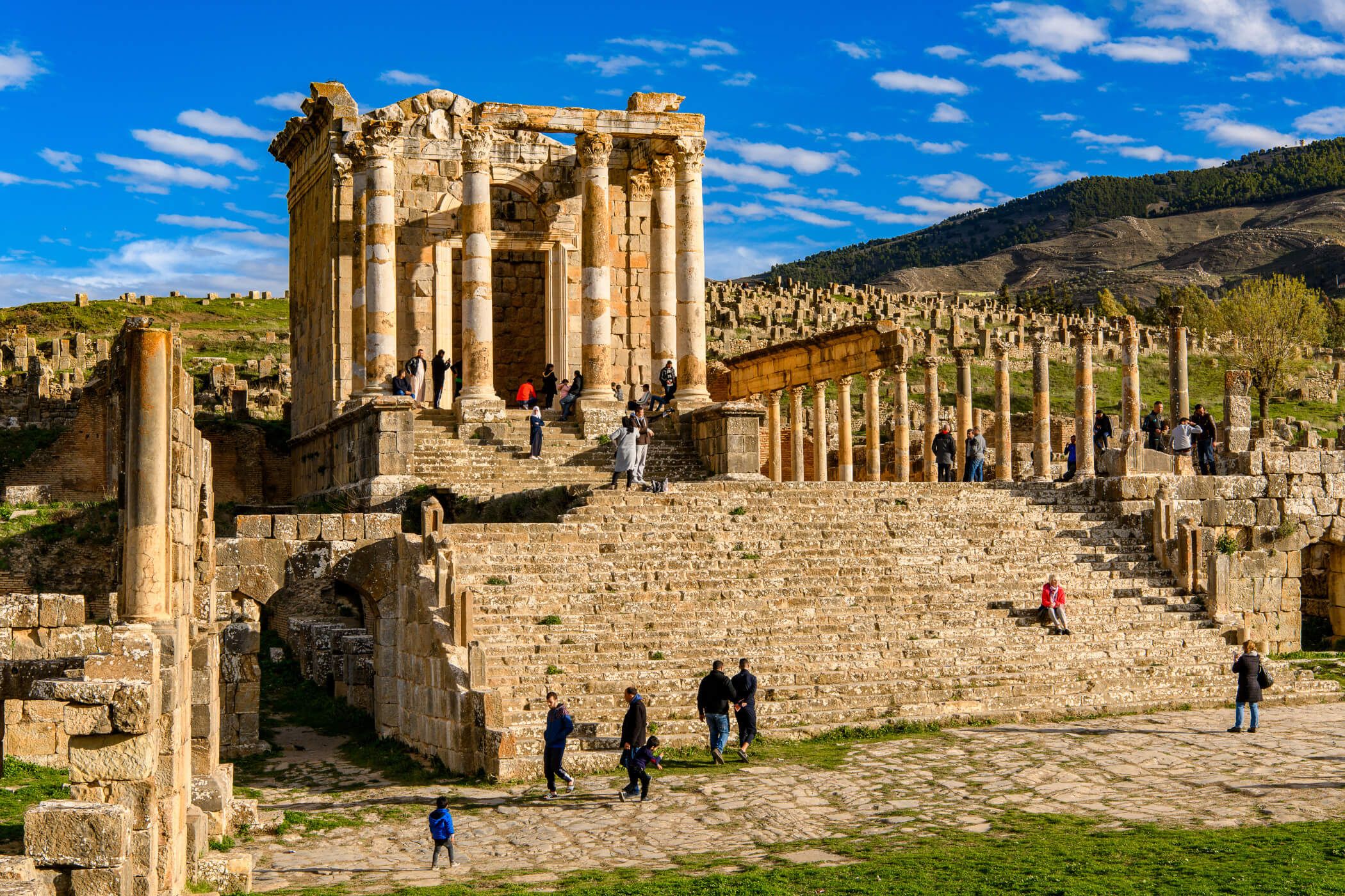 Ruinen mit Säulen und Treppen, umgeben von Besuchern und einer malerischen Landschaft unter blauem Himmel.
