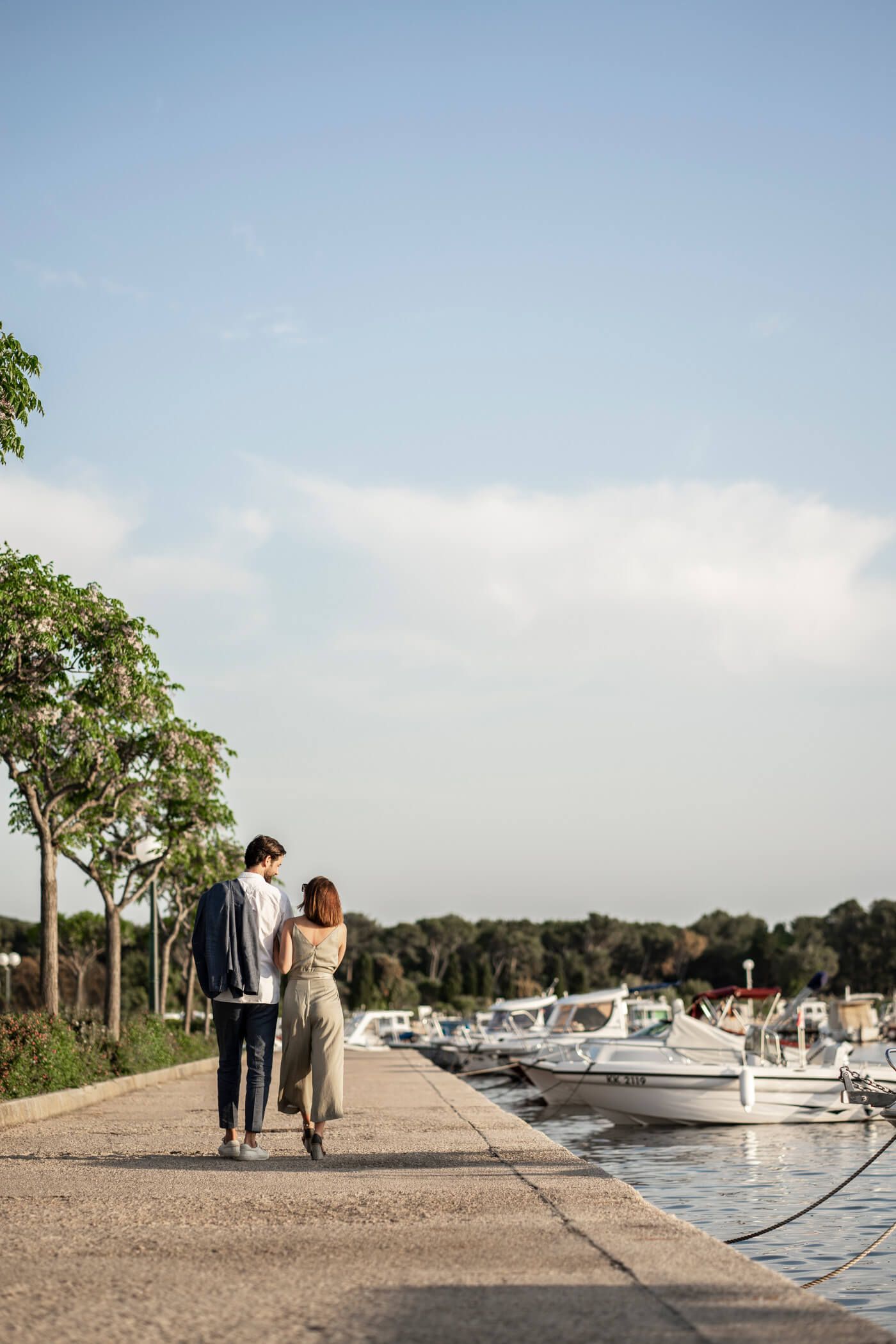 Ein Paar spaziert am Hafen entlang, umgeben von Bäumen und Booten, unter blauem Himmel.