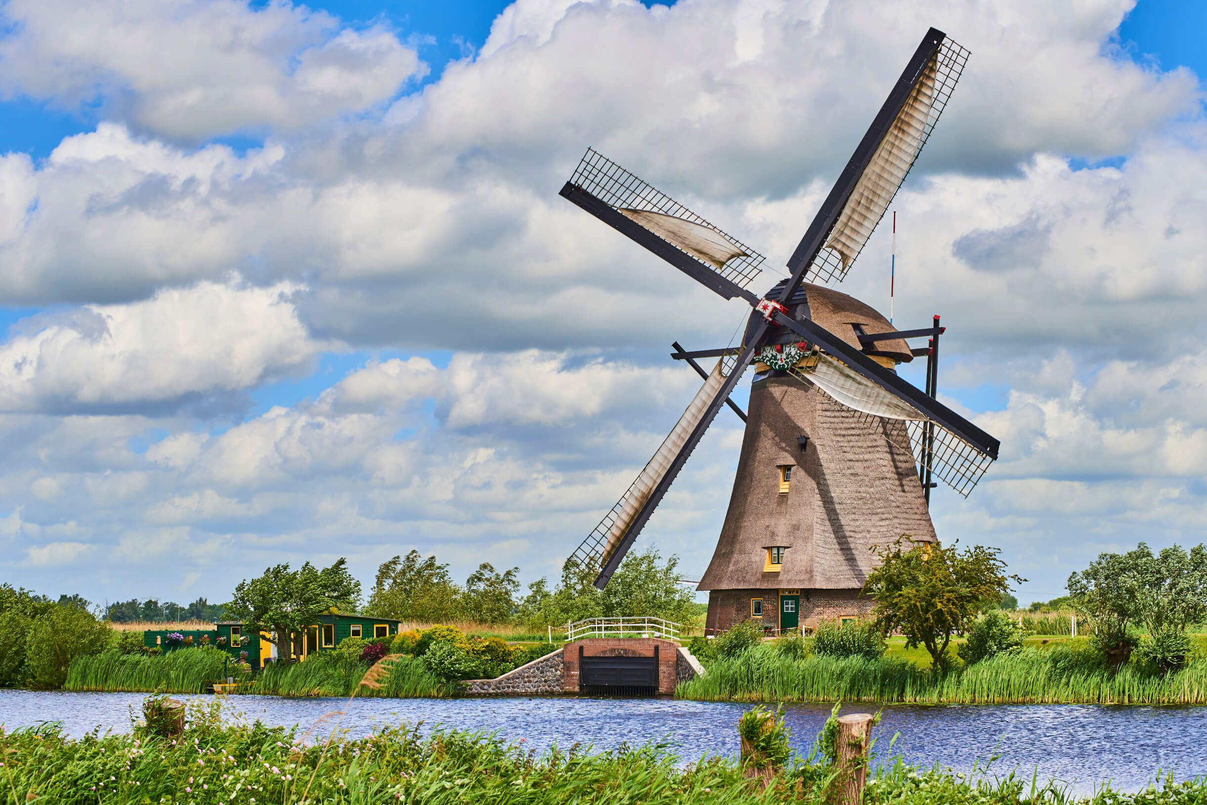 Eine Windmühle steht malerisch am Wasser, umgeben von grünem Gras und bewölktem Himmel.