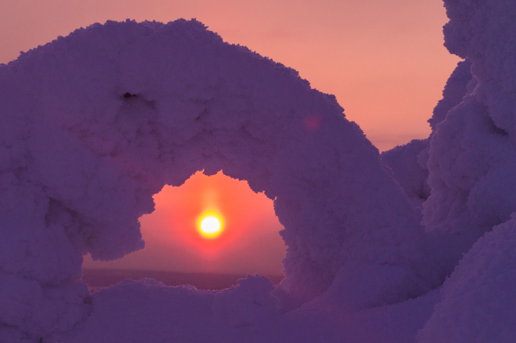 Eine schneebedeckte Struktur umrahmt die untergehende Sonne vor einem pastelrosa Himmel.