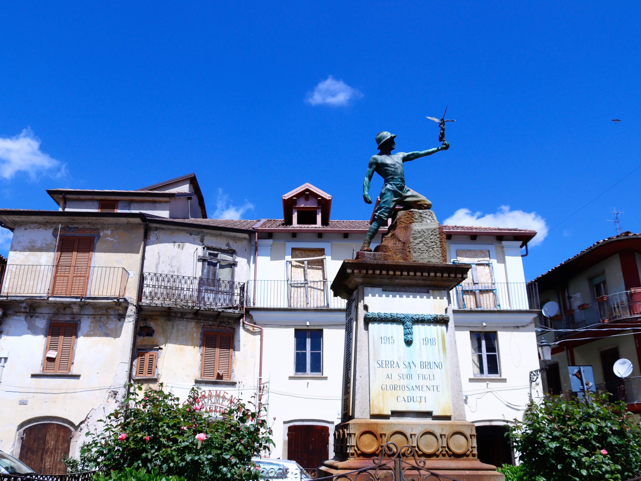Statue eines Soldaten, umgeben von alten Gebäuden und Blumen, unter blauem Himmel in Serra San Bruno.