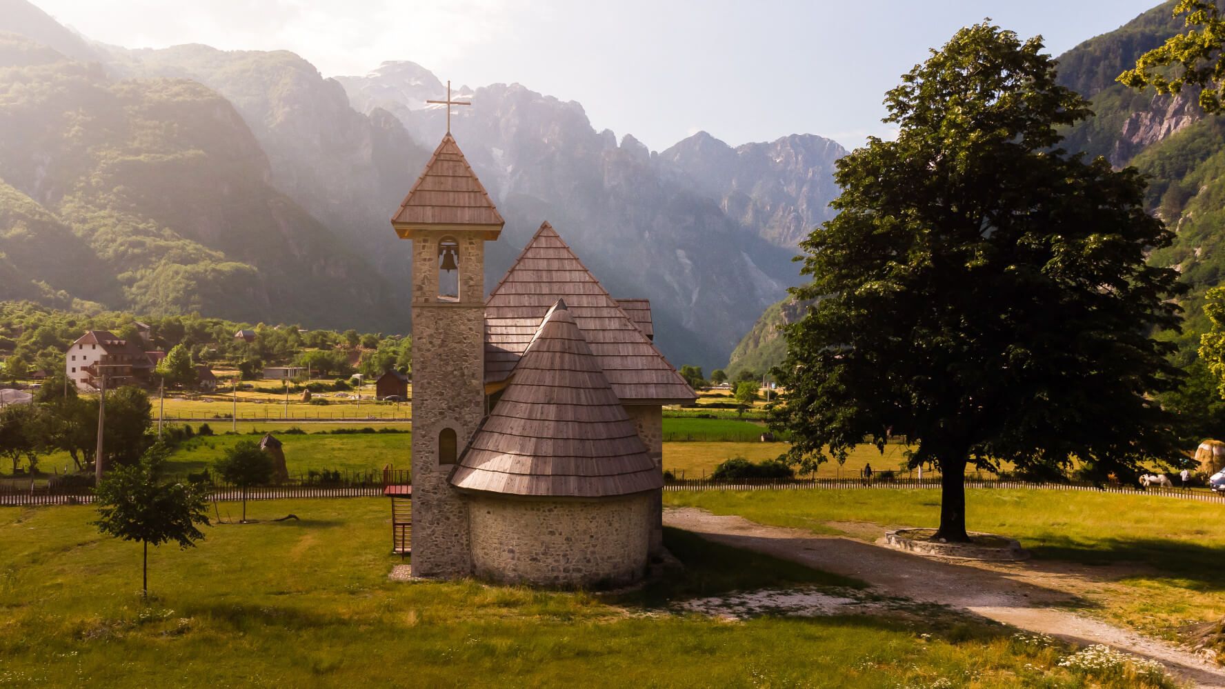 Eine malerische Kirche in einer grünen Landschaft, umgeben von hohen Bergen und einem großen Baum.