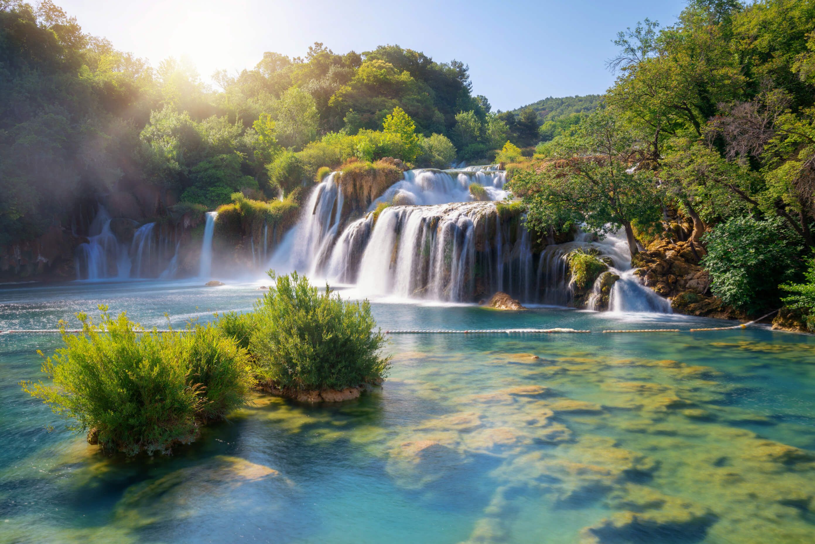 Ein malerischer Wasserfall im Nationalpark Plitvice  fließt sanft in einen klaren See, umgeben von üppigem Grün und sonnigem Himmel.