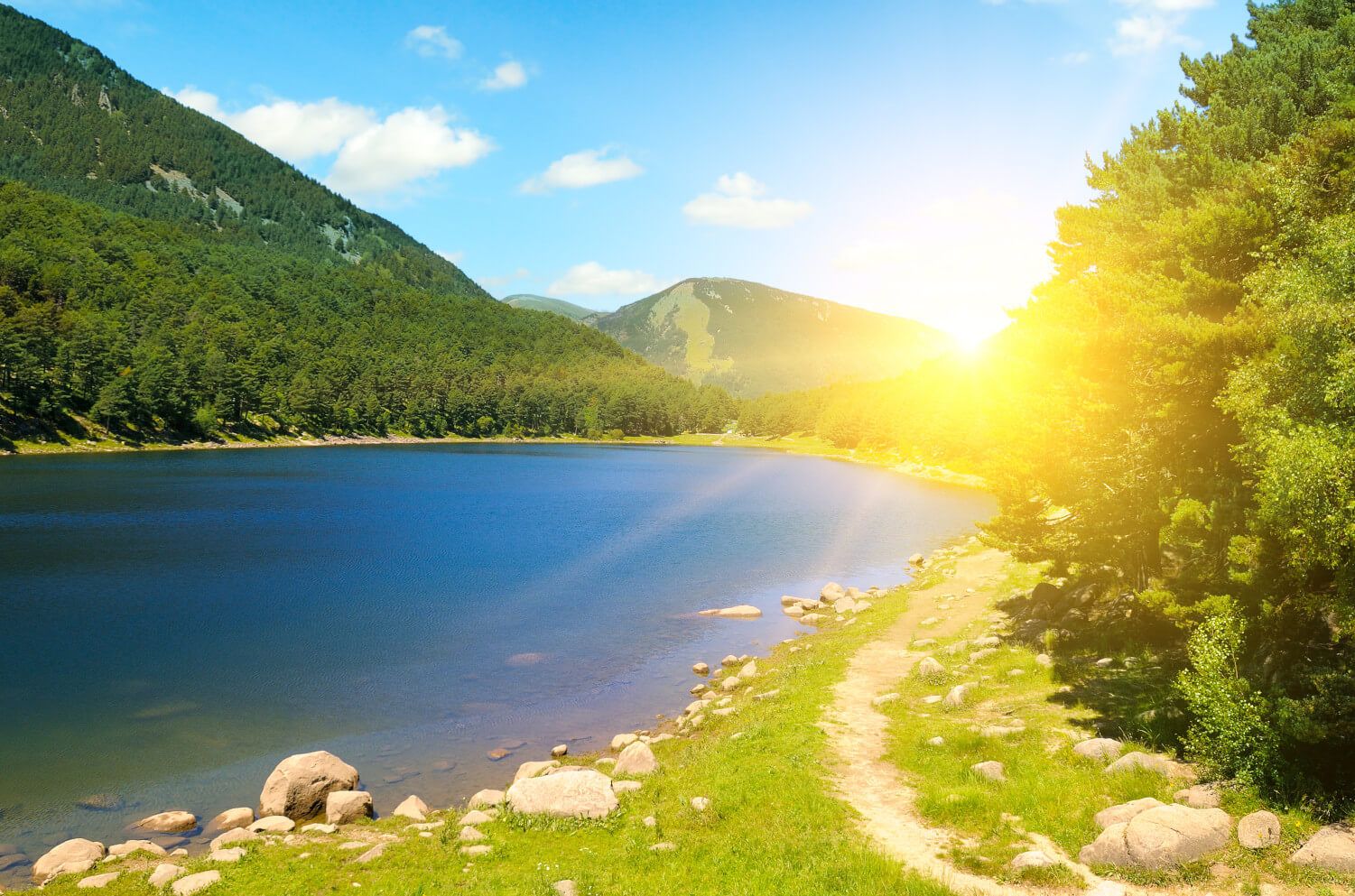 Ein ruhiger See umgeben von Bergen und Wäldern, mit strahlendem Sonnenlicht und blauen Himmel.