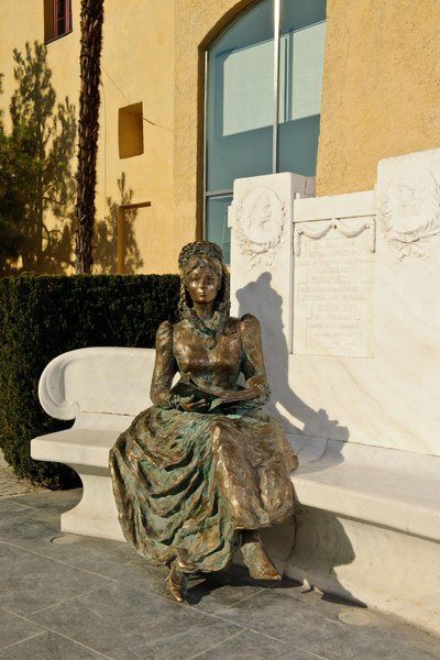 Eine Bronzeskulptur einer Dame in historischem Kleid sitzt auf einer marmornen Bank vor einer Wand.