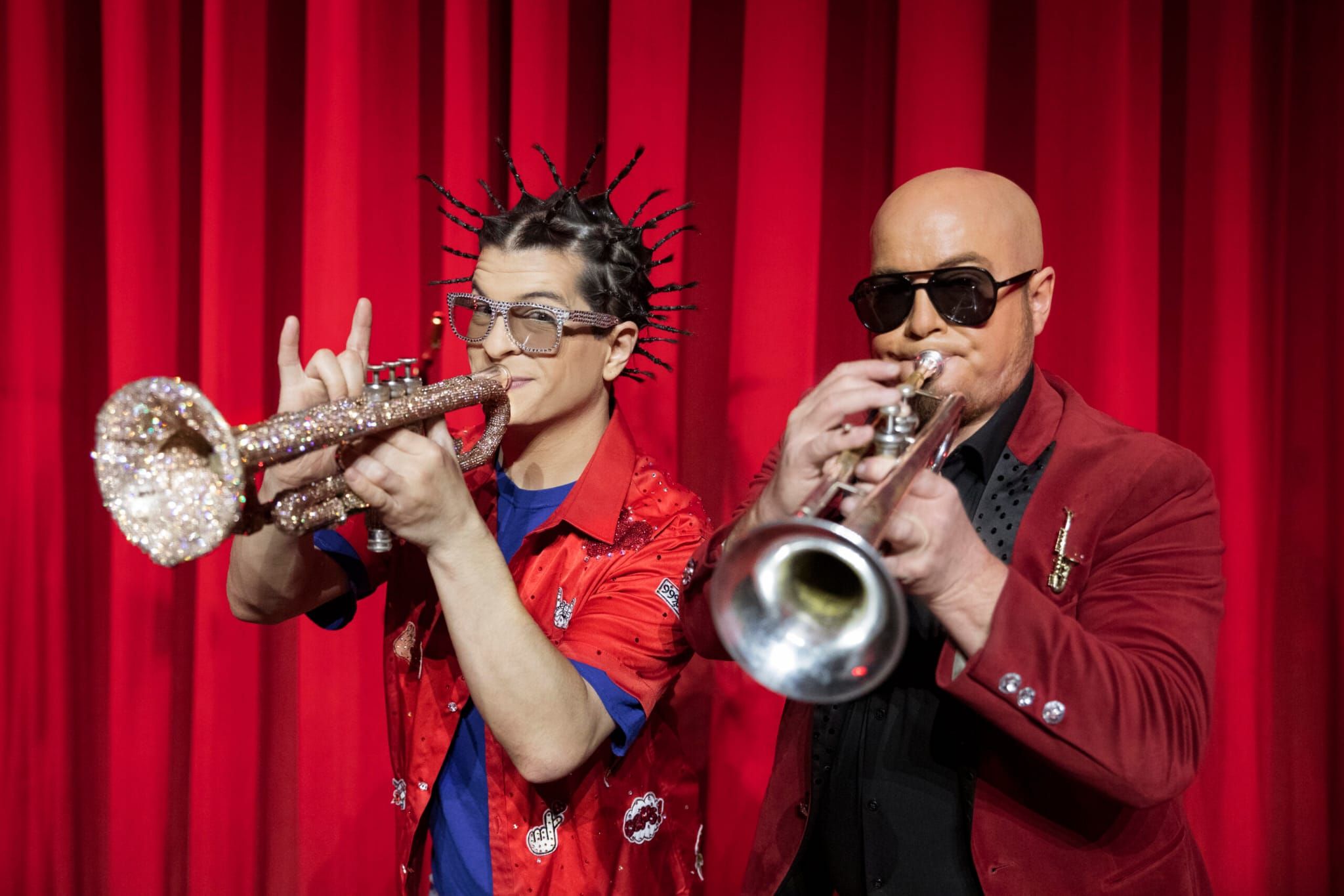 Zwei Musiker spielen Trompete, einer mit spitzen Haaren und Glitzertrumpet, der andere in Sonnenbrille und rotem Jackett.