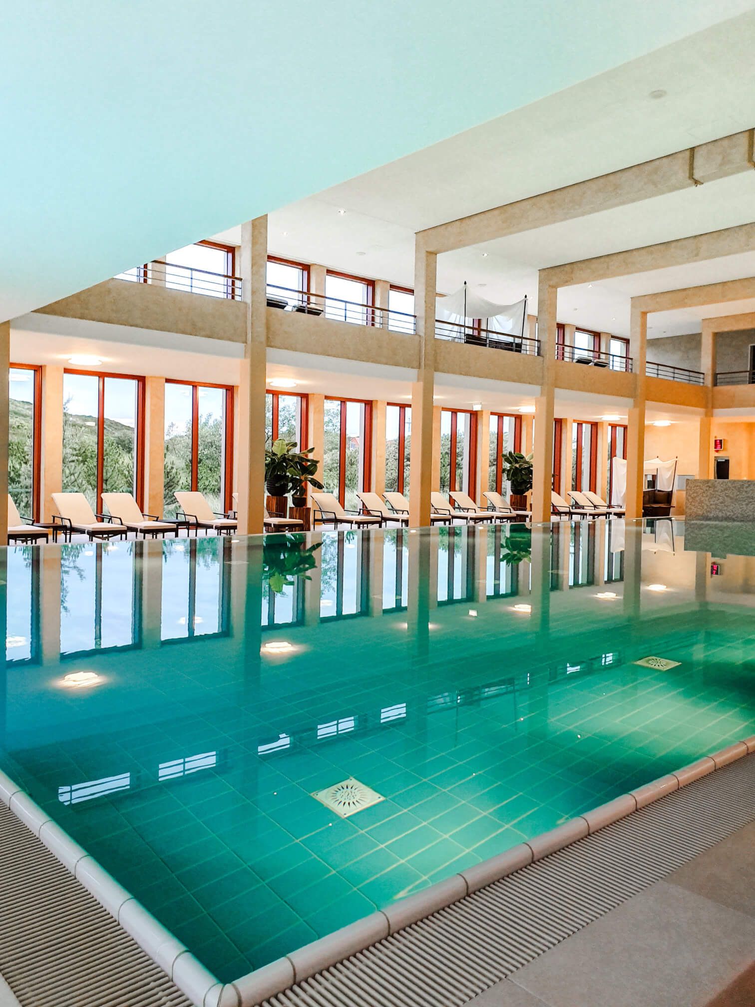 Ein modernes Hallenbad mit großen Fenstern, blauem Wasser und Liegen zum Entspannen. Helle, einladende Atmosphäre.