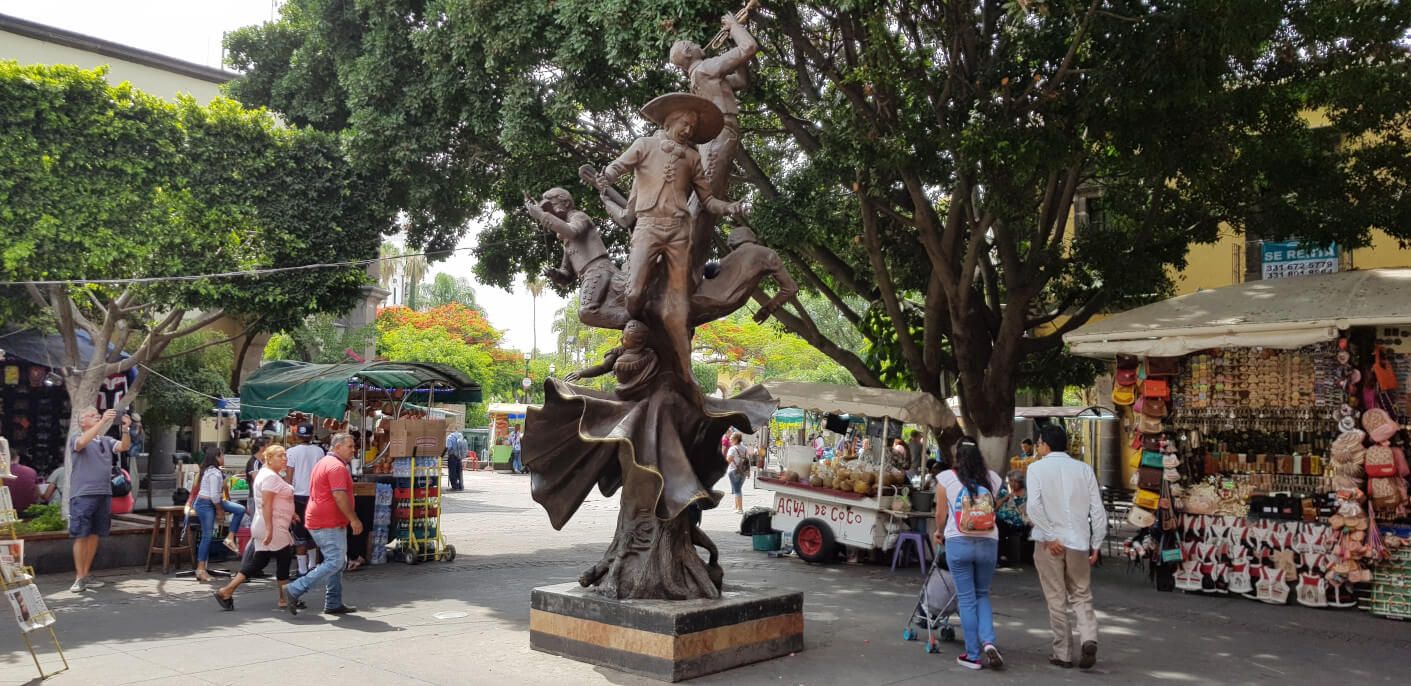 Eine lebendige Markt-Szene mit einer beeindruckenden Statue und vielen Menschen zwischen Verkaufsständen und Bäumen.