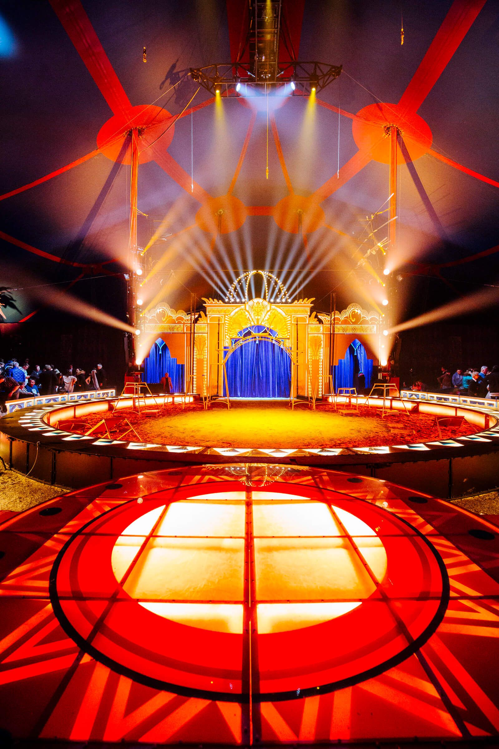 Die Zirkusmanege ist bunt beleuchtet, mit einem blauen Vorhang und festlichen Lichtern in einer großen Zeltatmosphäre.