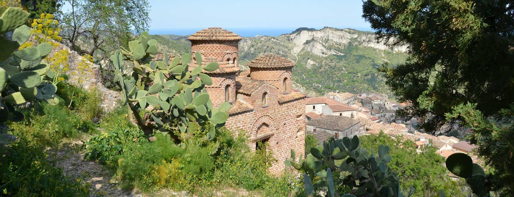 Eine malerische Landschaft mit alten Ziegelgebäuden, umgeben von grünem Bewuchs und Blick auf Meer und Berge.
