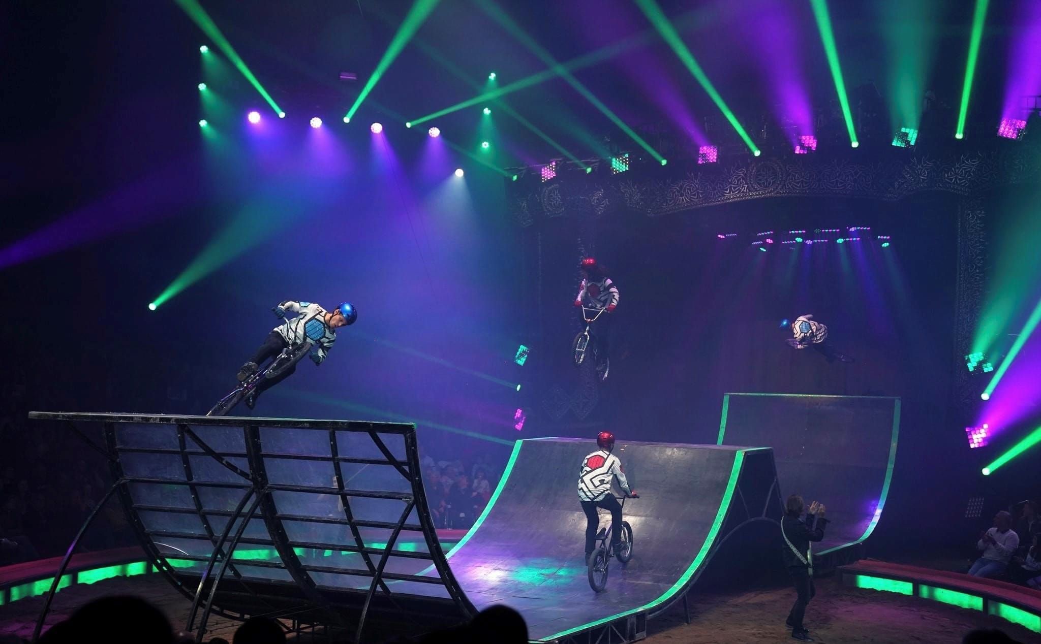 BMX-Fahrer zeigen spektakuläre Tricks in einer beleuchteten Arena, umgeben von jubelndem Publikum.