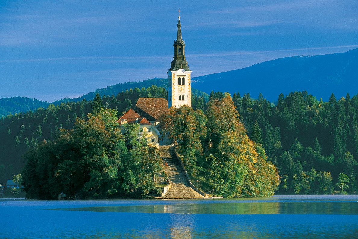 Eine malerische Insel mit einer Kirche, umgeben von Bäumen, am Ufer eines ruhigen Sees im Hintergrund Berge.