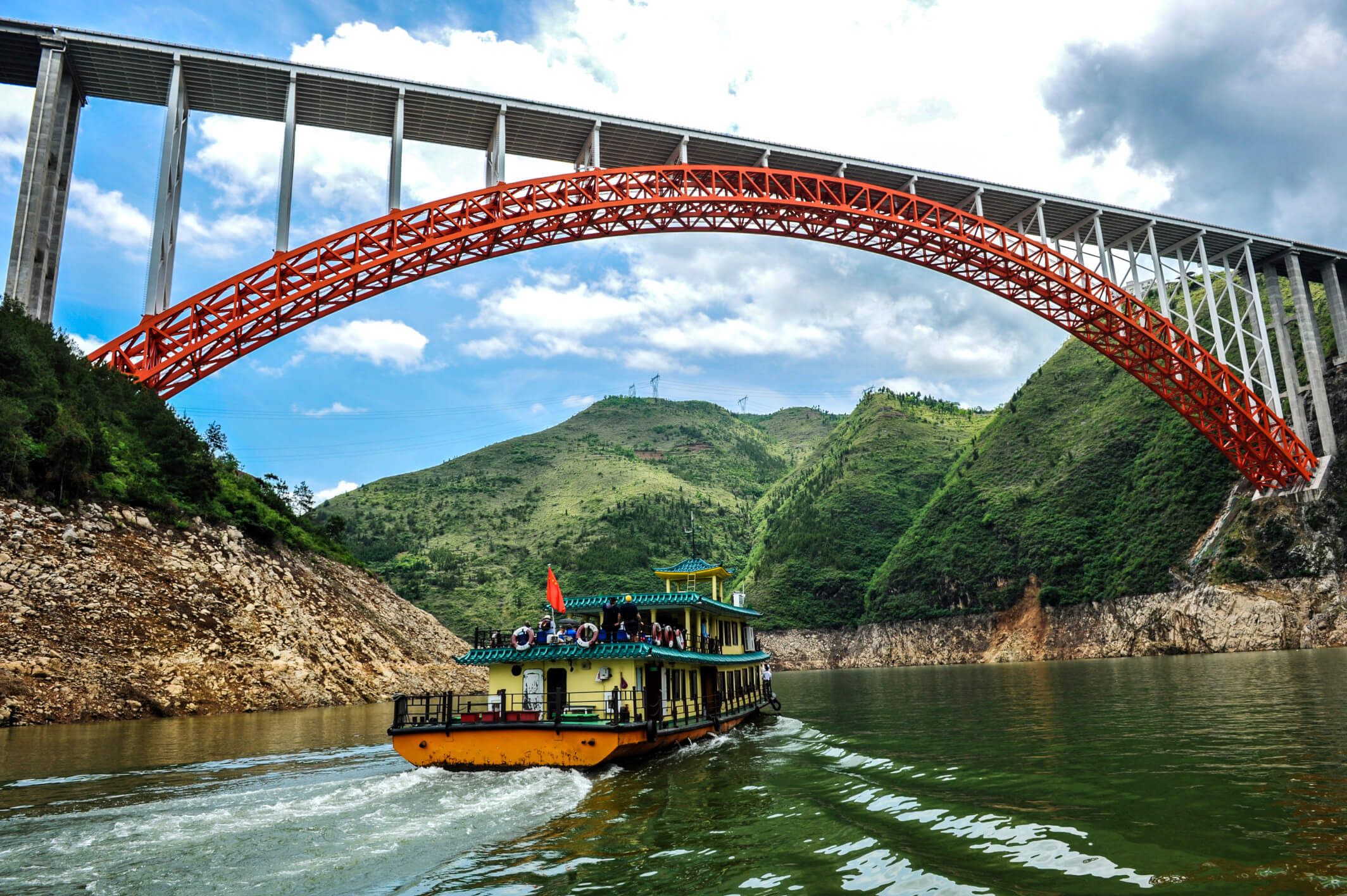 Ein buntes Boot fährt unter einer beeindruckenden, roten Brücke inmitten grüner Berge und blauem Himmel.