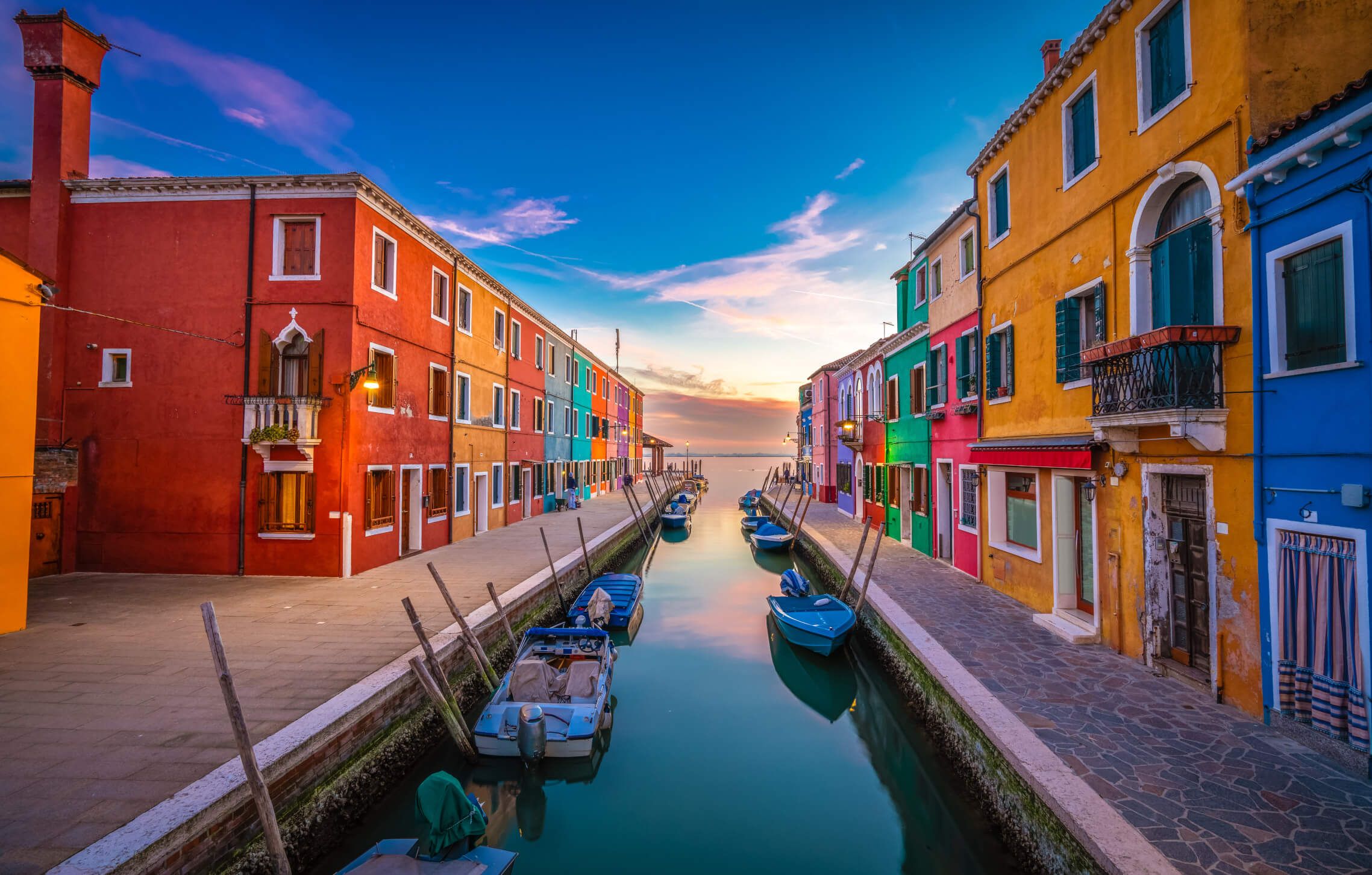 Bunte Häuser säumen einen ruhigen Kanal bei Dämmerung, mit Booten im Wasser und einem klaren Himmel.