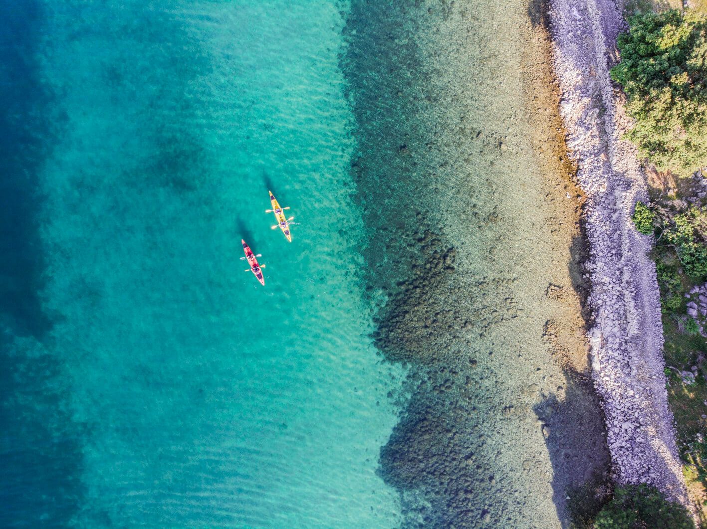 Zwei Kajaks paddeln im klaren Wasser, umgeben von einem Kiesstrand und üppiger Vegetation.