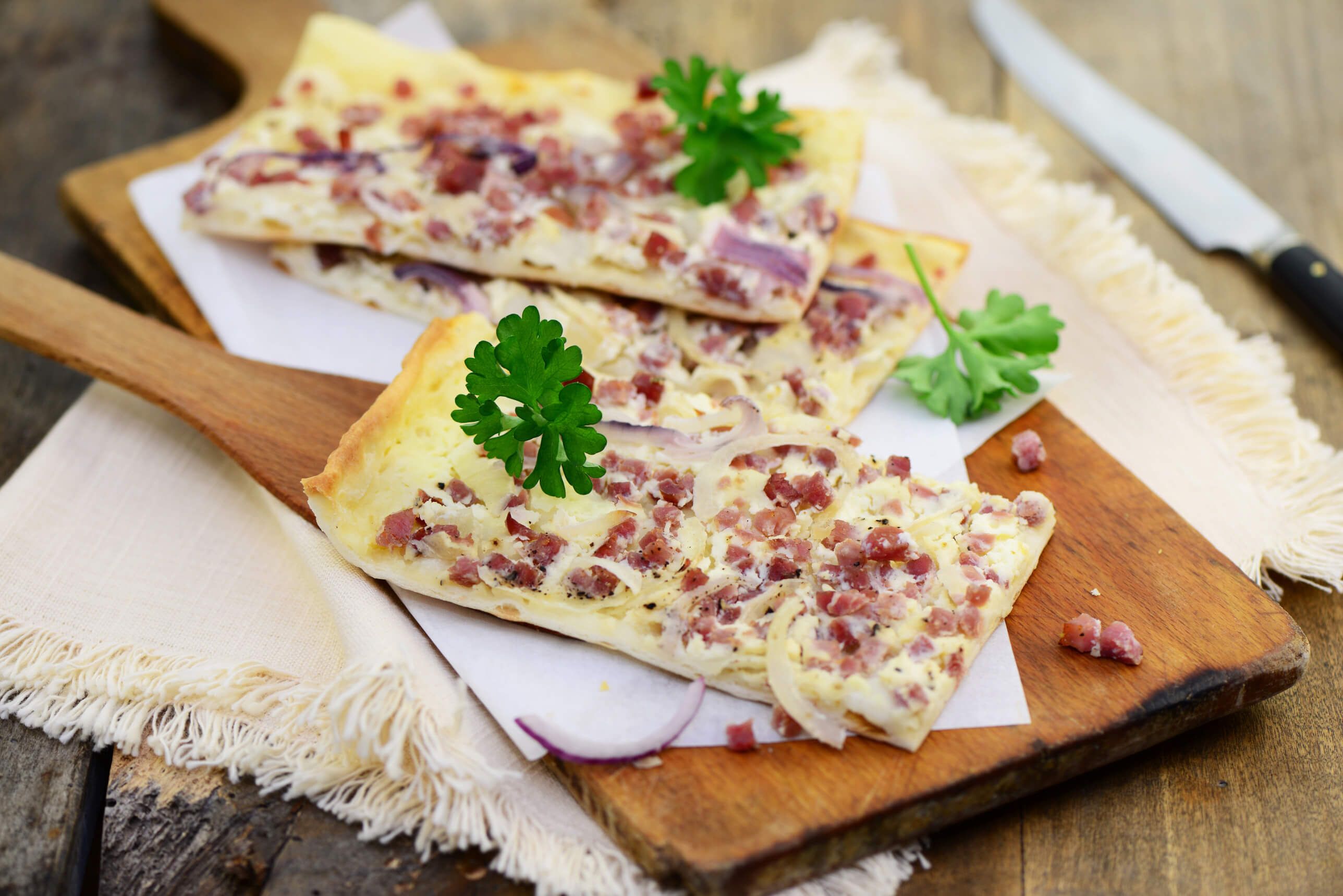 Leckere Flammkuchenstücke mit Schinken und Zwiebeln, garniert mit frischer Petersilie auf einem Holzbrett.