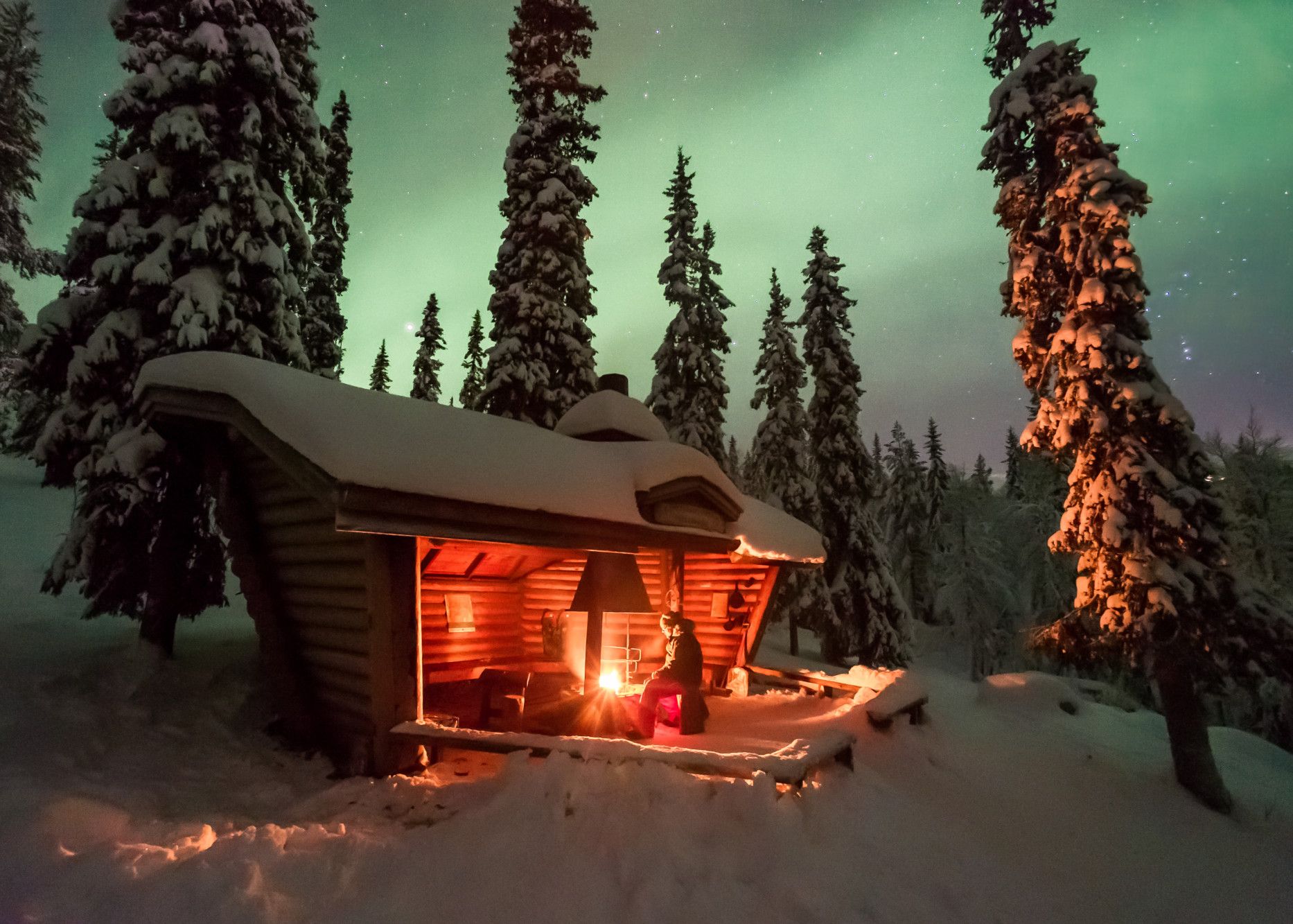 Eine gemütliche Hütte im verschneiten Wald, umgeben von Tannen und erleuchtet von geheimnisvollem Nordlicht.