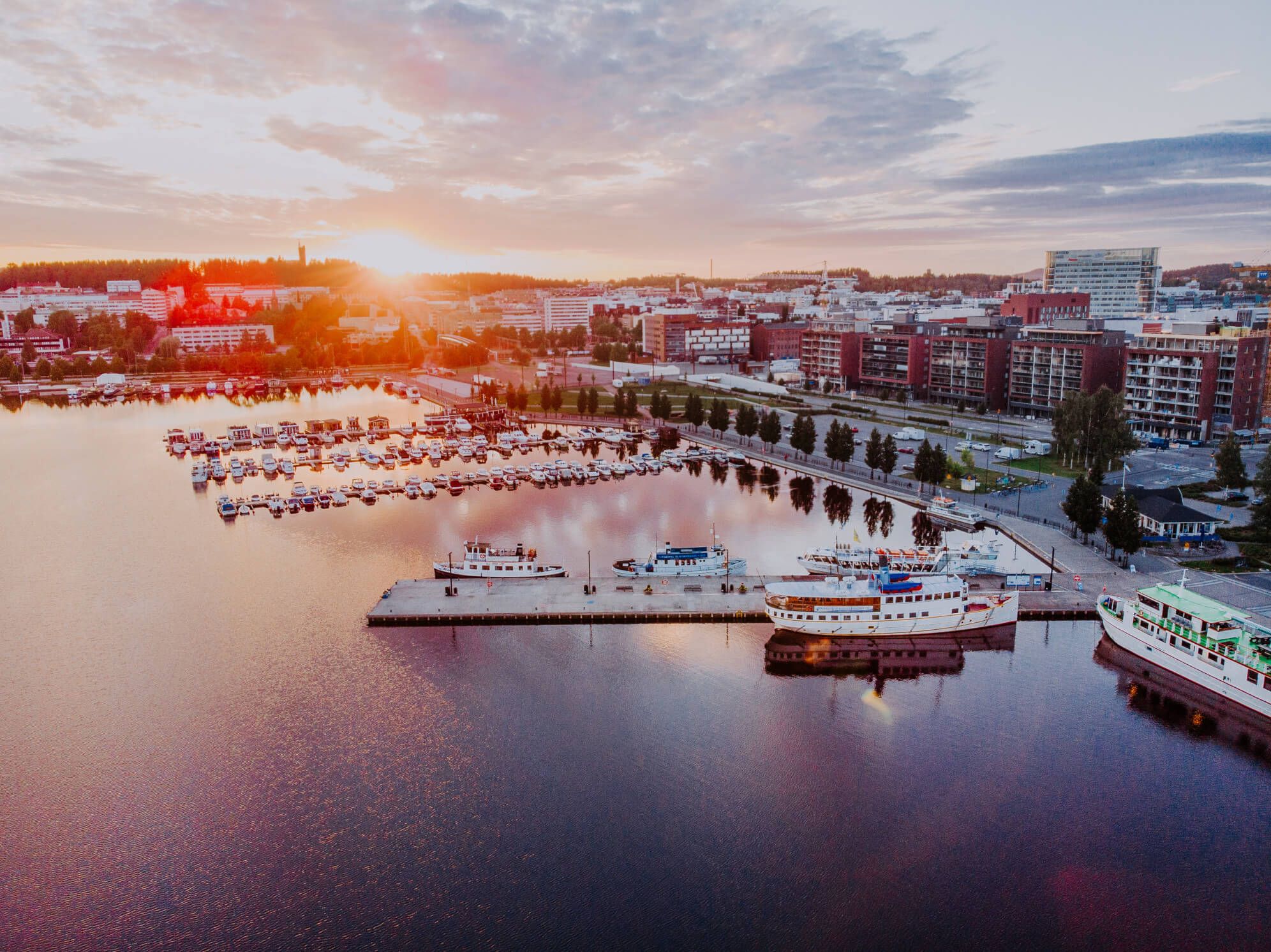 Sonnenuntergang über einem ruhigen Hafen mit Booten und Stadtlandschaft im Hintergrund. Reflektionen im Wasser.