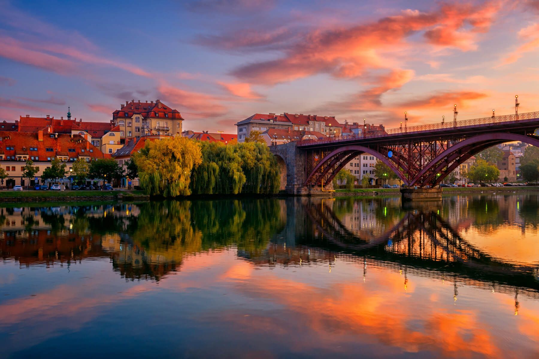 Eine malerische Brücke spiegelt sich im ruhigen Wasser bei Sonnenuntergang, umgeben von bunten Gebäuden und Bäumen.