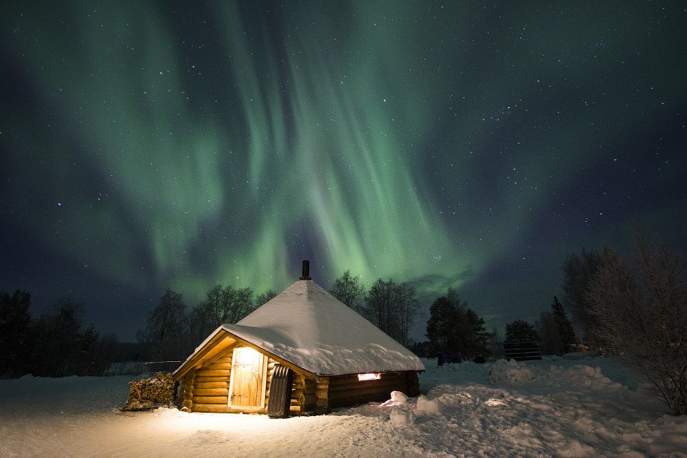 Eine gemütliche Holzhütte im Schnee, erleuchtet von Polarlichtern am nachtblauen Himmel.
