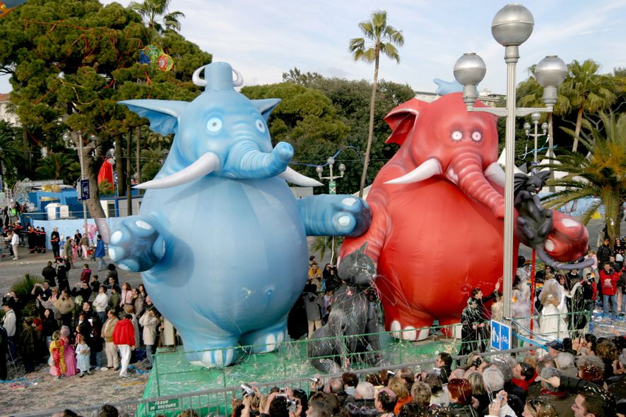 Riesenballons von blauen und roten Elefanten stehen neben einer Menschenmenge bei einem Fest, umgeben von Palmen.