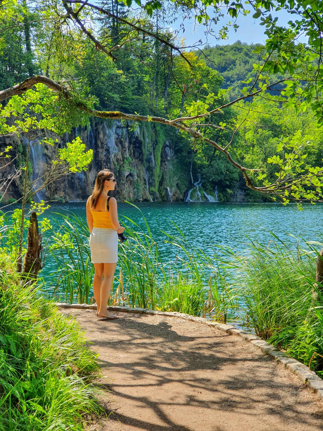 Eine Frau steht am Ufer eines klaren Sees im Nationalpark Plitvicer Seen , umgeben von üppigem Grün und sanften Wasserfällen im Hintergrund.