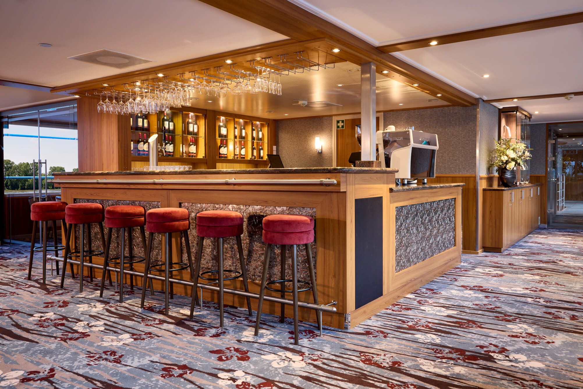 Eine elegante Bar mit Holzmöbeln, roten Hockern und Glasregalen für Weinflaschen erwartet die Gäste.