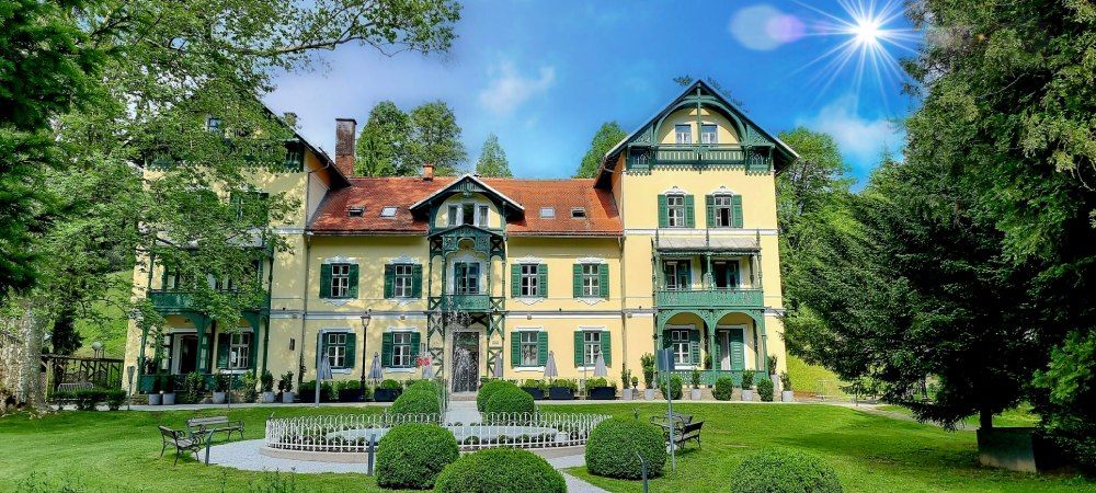 Eine elegante Villa mit grünem Garten, umgeben von Bäumen, unter blauem Himmel und strahlender Sonne.