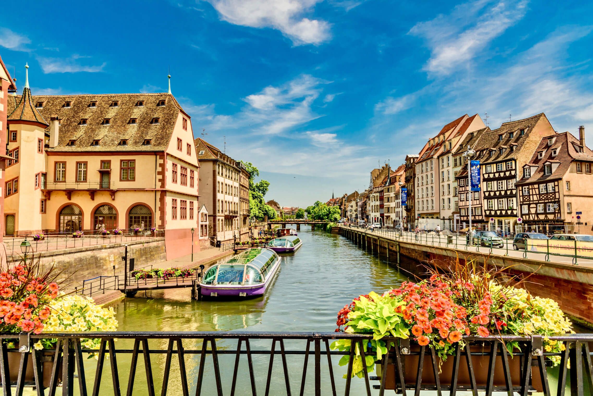 Blick auf einen Kanal mit bunten Booten, historischen Gebäuden und Blumen im Vordergrund unter blauem Himmel.